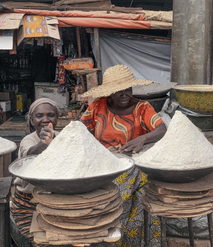 Two African women in traditional attire selling flour at a market stall.