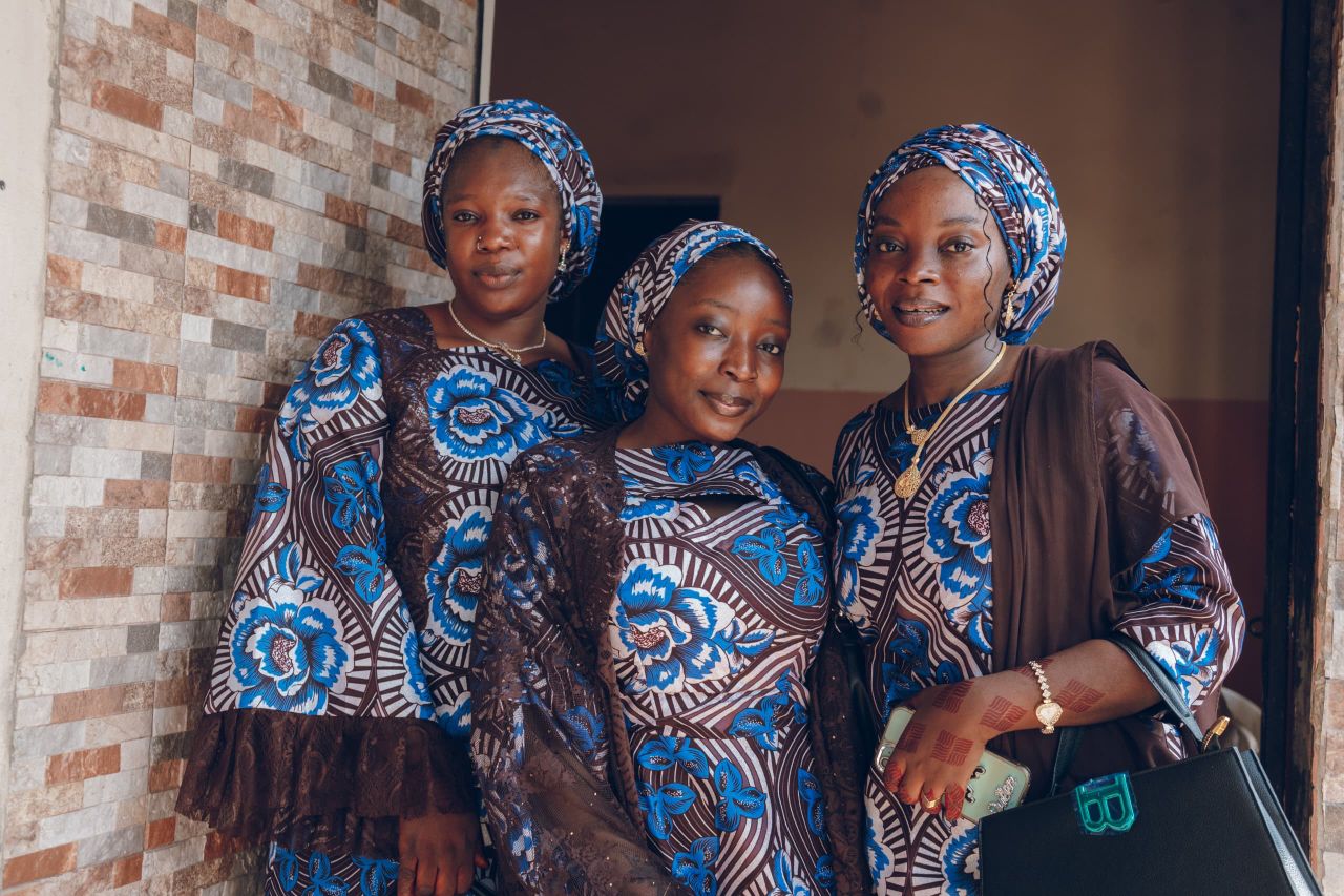Three women wearing elegant traditional African dresses pose indoors.