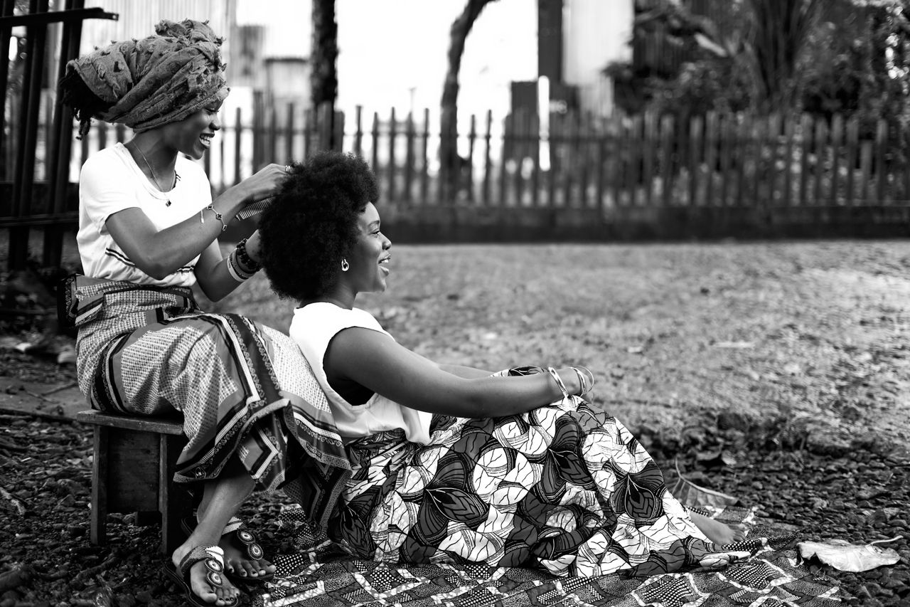 Black and white photo of two women in traditional attire, one braiding the other's hair.