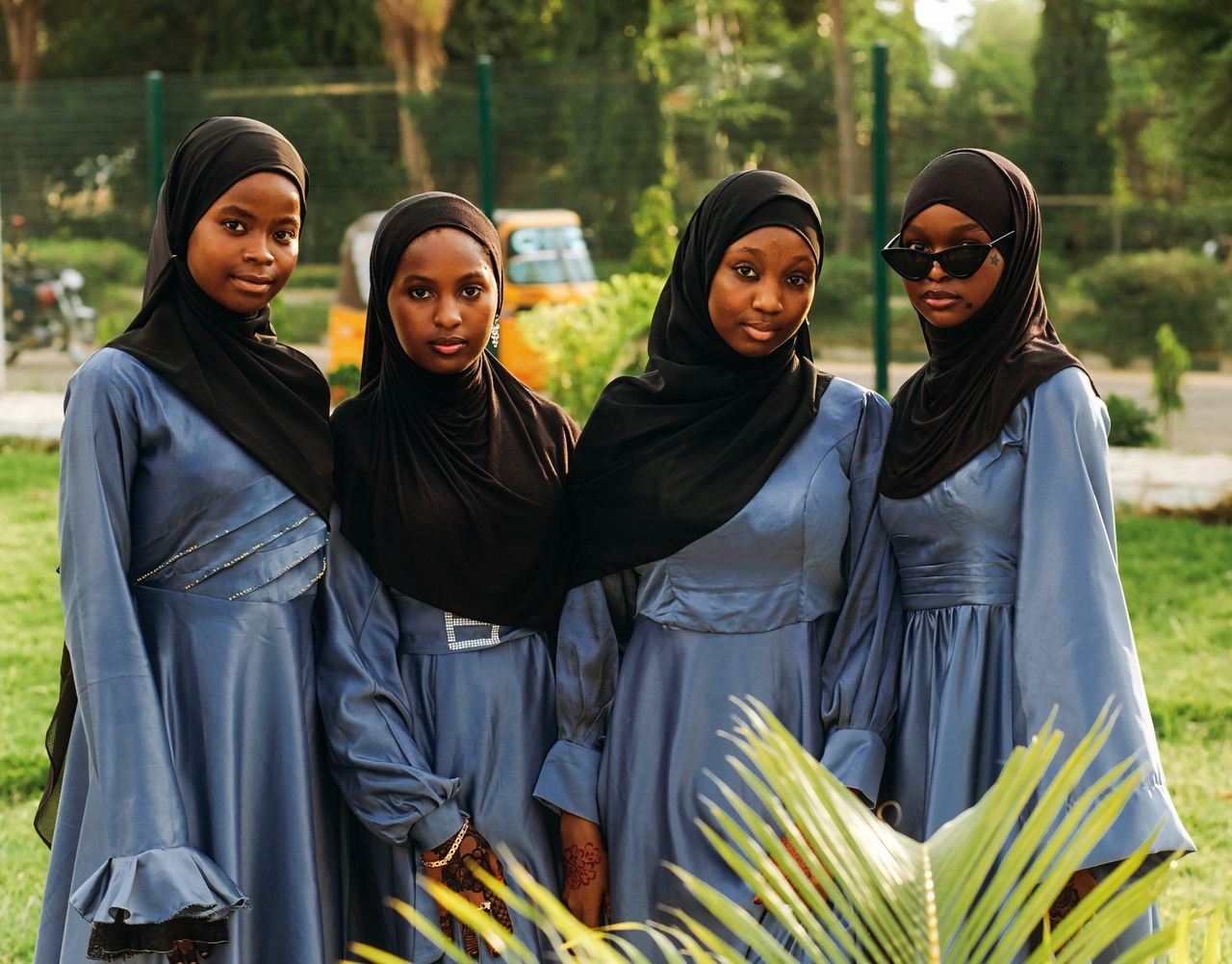 Four young women wearing hijabs and elegant dresses pose together in a sunny outdoor setting.